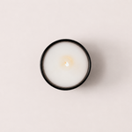 Load image into Gallery viewer, A top-down view of a lit white candle in a minimalist black-rimmed glass container, centered on a solid pale pink background.
