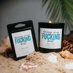 Load image into Gallery viewer, A candle and wax melt set with black packaging and white labels reading 'doing my FUCKING best' and 'HIGH TIDE', styled on sand with seashells and green leaves in the background.
