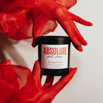 Load image into Gallery viewer, A black jar candle labeled 'ABSOLUTE SHIT SHOW' is held by red gloves wrapped in sheer red fabric against a plain light background.
