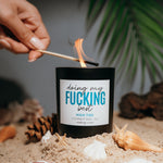 Load image into Gallery viewer, A black candle jar labeled 'doing my FUCKING best HIGH TIDE COCONUT WAX  CANDLE sits in sand with seashells around it, while a hand holds a lit match above it. Palm leaves and a dark background are visible behind.
