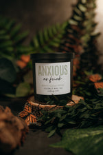 Load image into Gallery viewer, A black glass candle jar with a white label reading 'ANXIOUS as fuck' and 'WHITE EUCALYPTUS COCONUT WAX sits on a wood slice surrounded by green leaves, dried foliage, and a butterfly ornament.
