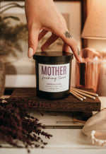 Load image into Gallery viewer, A hand with a tattoo reaches down to a black jar candle labeled 'MOTHER FUCKING TIRED' on a rustic wooden shelf, surrounded by cozy home decor props like cotton swabs, dried flowers, and a lamp in the background.
