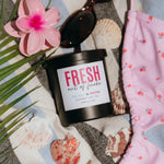 Load image into Gallery viewer, A black jar candle labeled 'FRESH out of fucks' is styled in a beach-themed flat lay with sunglasses, a pink plumeria flower, seashells, palm leaves, a pink floral bikini top, and a striped beach towel.
